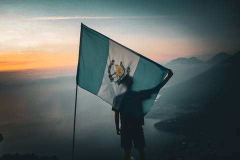       Silhouetted hiker holds Guatemalan flag aloft atop mountain ridge at colourful dawn.
  