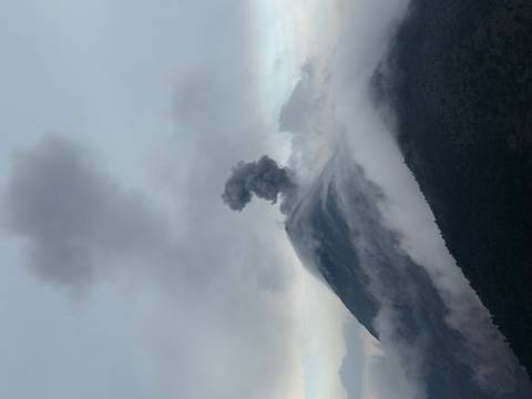       Volcano summit releasing thick plume of dark ash into cloudy sky with misty forested slopes below.
  