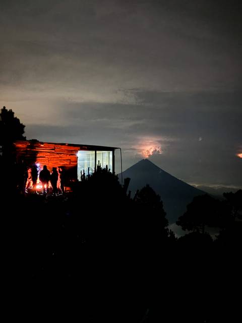      Silhouetted hikers warming by a fire outside a modern cabin overlooking a conical volcano glowing in the night sky
  
