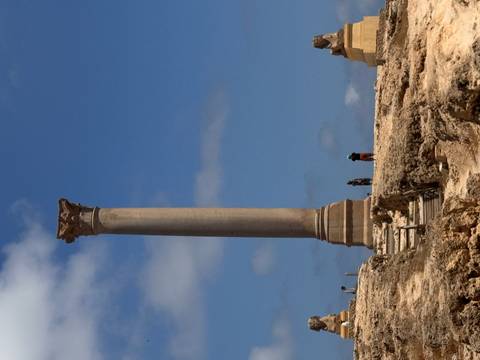       Travelers stand dwarfed by a towering solitary column against deep blue sky on desert ruins
  