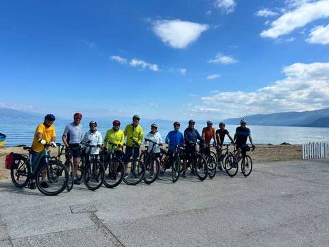       Cycling group lined up with touring bikes beside a calm lakeshore under blue skies.
  