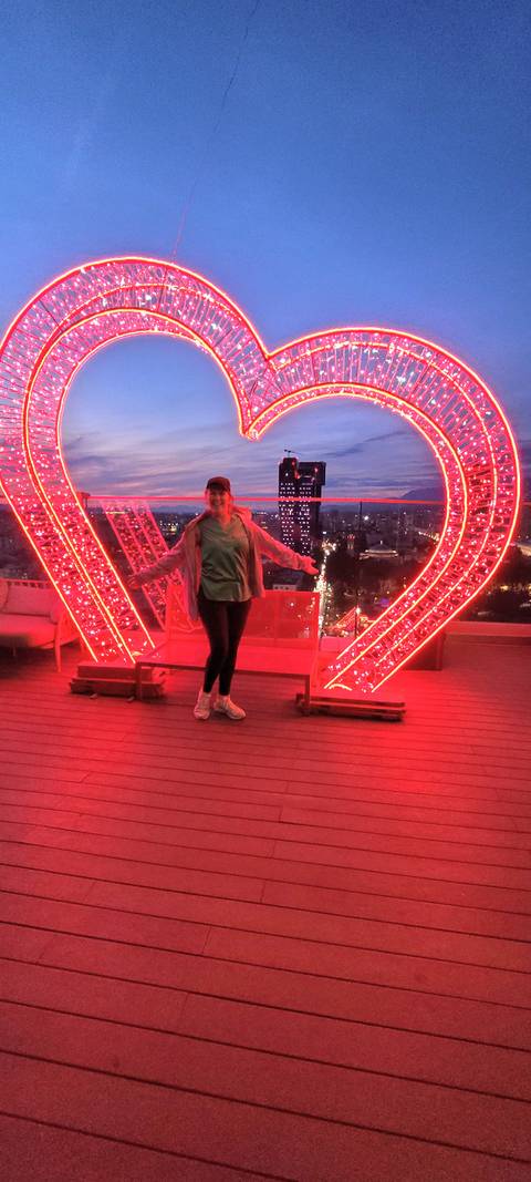       Smiling traveller stands under bright red neon rooftop installation overlooking a city at dusk
  