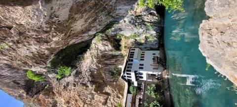       White Dervish monastery tucked beneath a towering cliff beside teal spring waters at Blagaj Tekke
  