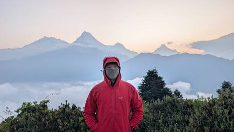       Trekker in red jacket stands before misty Himalayan range at dawn, clouds swirling below.
  