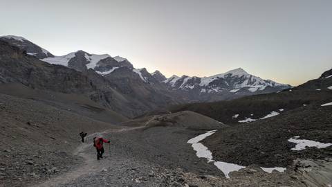       Trekkers navigate a rocky alpine path at dawn with towering snowy peaks ahead.
  