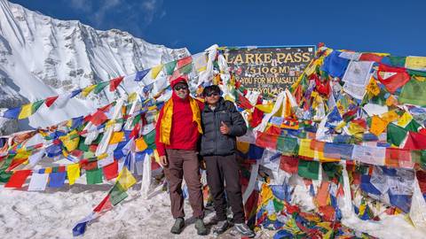       Two trekkers pose at snowy Larke Pass sign surrounded by colorful prayer flags at 5106 m.
  