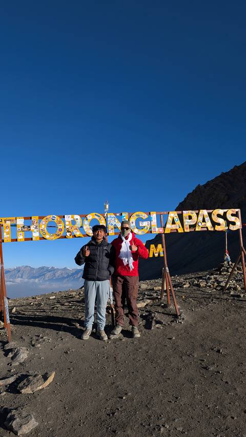       Two trekkers give thumbs-up beneath the colorful THORONG LA PASS summit sign against clear blue sky.
  