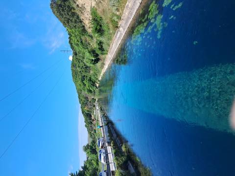       A crystal-clear blue spring forms a calm channel beside a rural roadside under a bright sky.
  