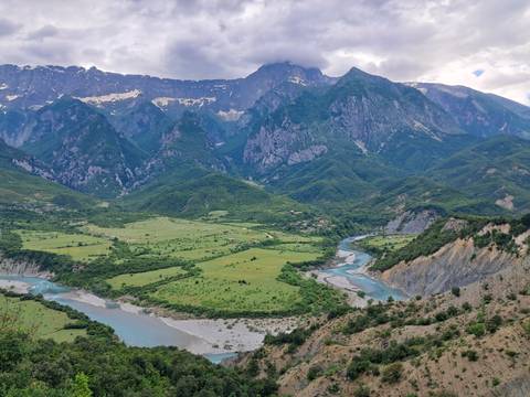       A sweeping alpine valley with a winding blue river sits beneath towering green mountains capped with lingering snow.
  
