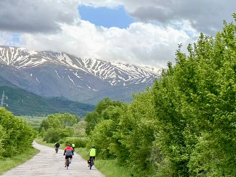       Cyclists ride a rural road lined with lush trees toward snow-capped Albanian mountains.
  