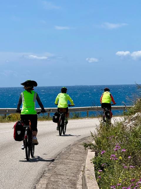       Three cyclists in neon vests ride along a seaside road beside deep blue water.
  