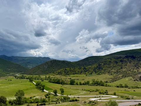       Wide Albanian valley with patchwork fields and forested hills under dramatic clouds.
  