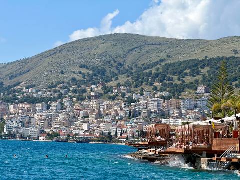       Seaside city of Saranda with terraced white buildings climbing a green hillside beside bright blue water and wooden sun decks.
  