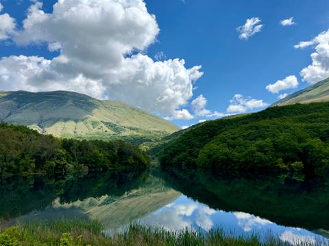       Mirror-like lake reflecting lush green hills and billowing clouds beneath a blue sky.
  