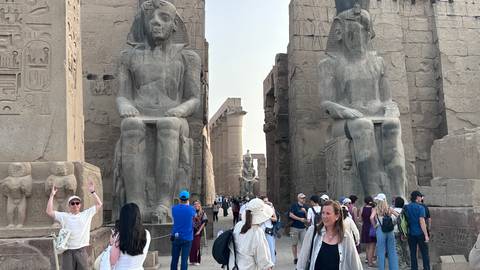       Crowds walk between towering seated statues at Luxor Temple during daylight
  