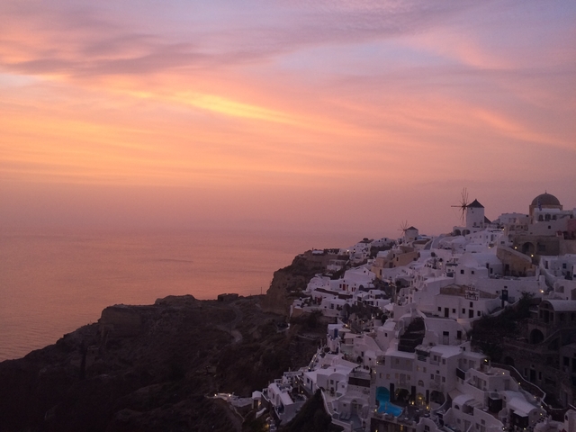       White buildings with blue domes on a cliffside overlooking a pink sunset.
  