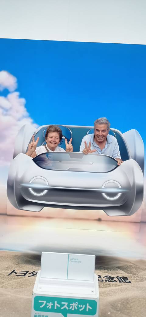       Smiling senior couple flashing peace signs while seated in a futuristic concept car display backdrop.
  