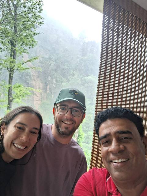       Close selfie of three travellers on a misty day with blurred green cliffs behind a bamboo screen.
  