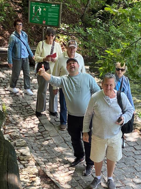       Tour group walking along cobbled path through shaded woodland, one man giving a thumbs up.
  