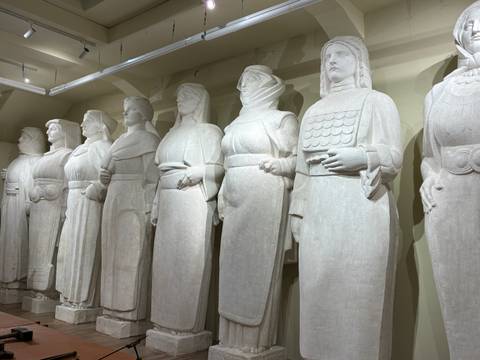       Row of life-size white stone statues of women in traditional dress displayed in a well-lit hall.
  