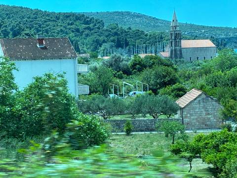       Motion-blurred view from a bus window of Croatian countryside with a church tower and olive trees.
  