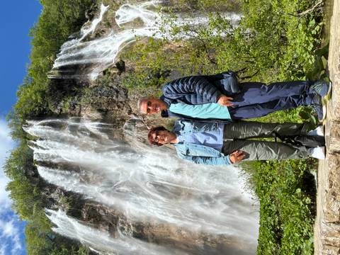       Couple stand smiling before a towering multi-tiered waterfall surrounded by lush greenery at Plitvice.
  