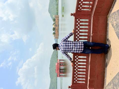       Man in plaid shirt leans on a red railing looking across Jal Mahal Lake toward the palace and green hills.
  