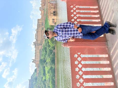       Traveler poses by the lakeside railing with Amber Fort rising on the opposite hill under a blue sky.
  