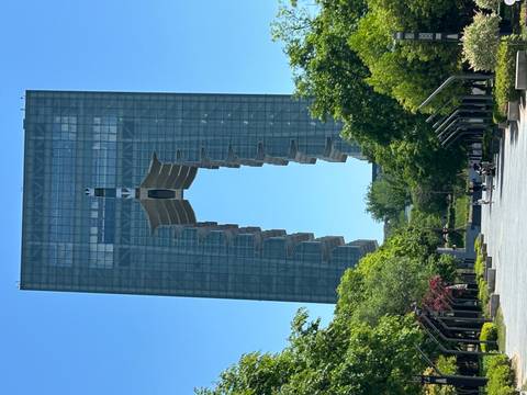       Modern glass arch building with large open portal framed by green trees and small people below
  