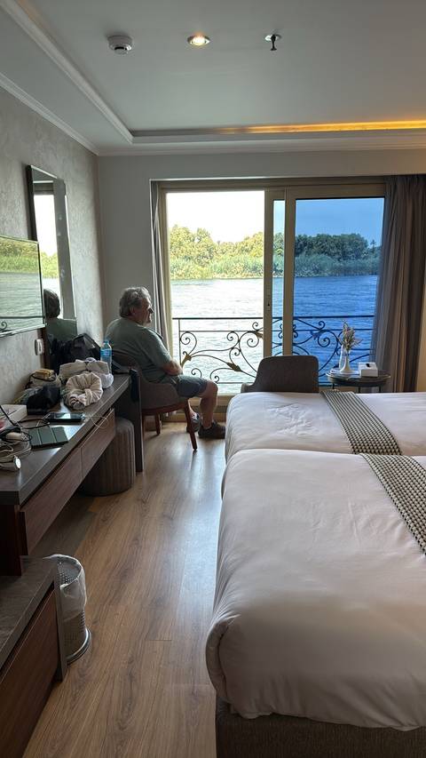      Older man relaxing in a river-view cruise cabin, seated by a window overlooking the Nile.
  