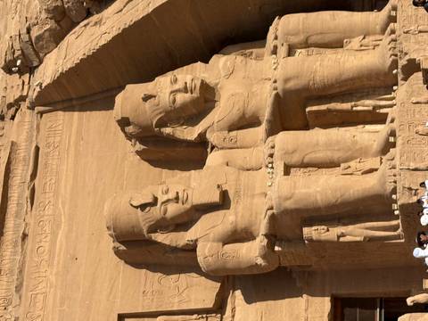       Gigantic seated statues of Ramses II carved into the sandstone cliff at Abu Simbel with tourists below for scale.
  