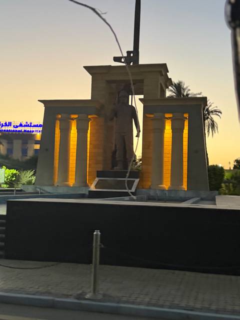       Blurry low-light photo of an illuminated statue between two columns beside neon Arabic signage.
  