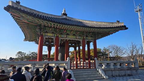       Traditional Korean bell pavilion with painted eaves stands in open courtyard as visitors gather.
  