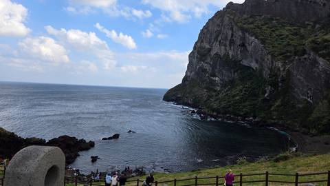       Volcanic cliff and bay on Jeju Island with visitors gathered along the shoreline.
  