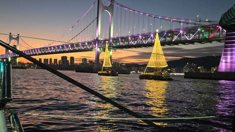       Color-lit Gwangan Bridge and Christmas tree boats shimmer at dusk over Busan bay.
  