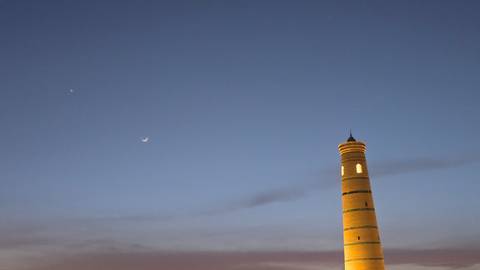       Slim illuminated minaret reaches into a deep twilight sky beside a crescent moon.
  