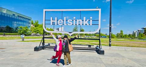       Two smiling women posing under a large outdoor Helsinki sign on sunny day.
  