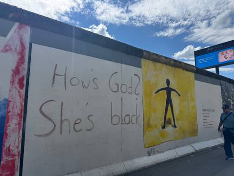       Section of Berlin Wall art reading 'How's God? She's black' with visitor.
  