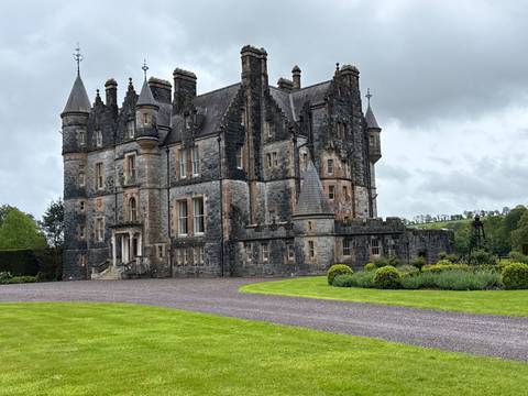      Grand gothic-style stone castle with turrets and manicured lawn under grey clouds.
  