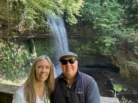       Smiling couple posing in front of a cascading forest waterfall surrounded by lush mossy rocks.
  