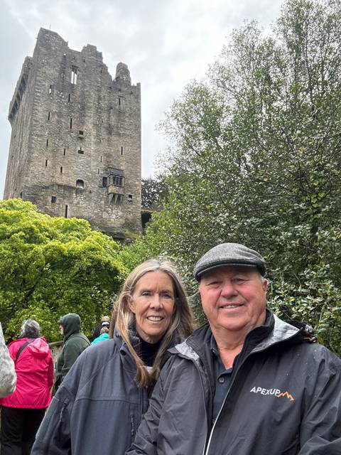       Couple standing beneath the tall stone tower of Blarney Castle amid leafy trees.
  