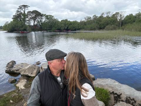       Couple sharing a kiss on rocky lakeshore with calm water and moored red boats in the background.
  