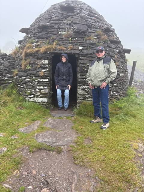       Two travelers at the doorway of an ancient beehive stone hut on a misty hillside.
  