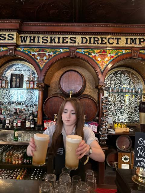       Bartender pouring a drink behind an ornate wooden bar with shelves of spirits.
  