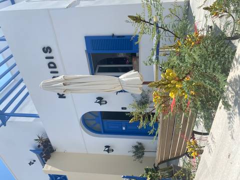       Charming whitewashed Greek building with blue door, shutters and pergola around a courtyard bench and plants.
  