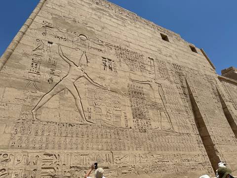       Exterior wall of an Egyptian temple carved with large-scale battle reliefs under bright sun.
  