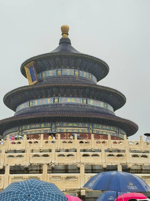       Close view of ornately tiled Temple of Heaven under gray sky with visitors carrying umbrellas
  