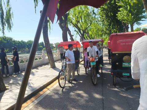       Rickshaws and cyclists moving along tree-lined canal in Beijing hutong
  