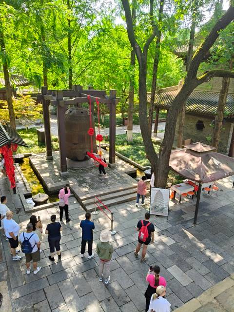       Visitors ringing a large bronze bell at a temple courtyard surrounded by trees
  