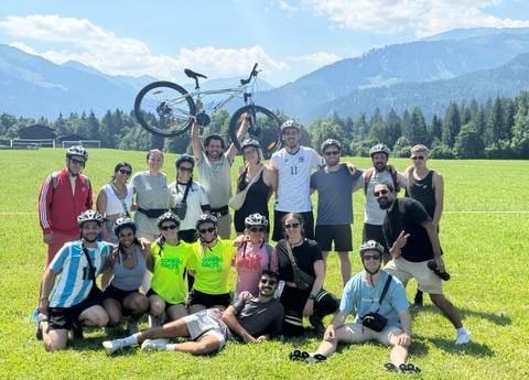       Large group of cyclists posing in a lush alpine meadow with mountains in the background, one lifting a bike overhead.
  